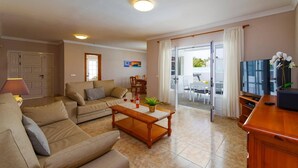 Ein Wohnzimmer mit einer Couch, einem Couchtisch und einem Fernseher. Eine Tür führt zu einem Balkon mit Tisch und Stühlen.