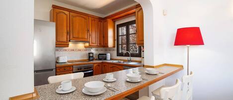 A compact kitchen with wooden cabinets, a granite countertop, a dining area with white chairs, and a red floor lamp.