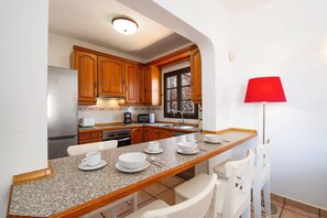 Cozinha compacta com armários de madeira, bancada de granito, área de refeições com cadeiras brancas e luminária de chão vermelha.