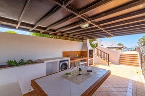 Uma área de cozinha externa com máquina de lavar embutida, uma mesa com copos de vinho e uma garrafa, e uma escadaria que leva a um andar superior.