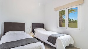 A bedroom with two beds, a window with a view of palm trees, and a bedside table with a lamp. - Villa Kikay by Villa Plus (Tías)