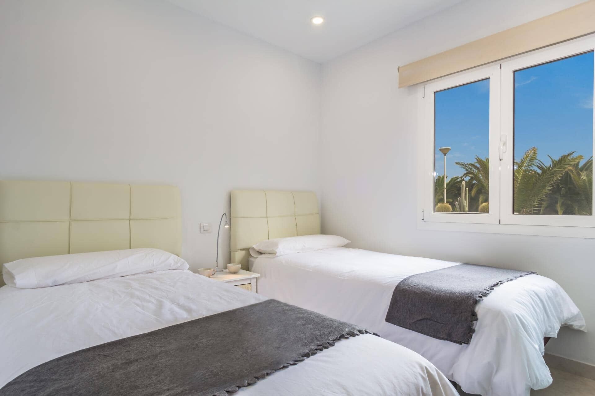 A bedroom with two beds, a nightstand, a lamp, and a window with a view of palm trees.