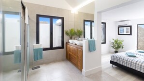 A modern bathroom with a glass shower, a wooden vanity, and a bed with a blue and white checkered duvet. - Villa JJ Beach by Villa Plus (Tías)
