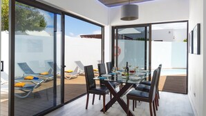 A modern dining area with a glass table and chairs, overlooking a pool and lounge area. - Villa JJ Beach by Villa Plus (Tías)