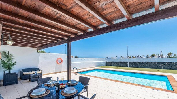 An outdoor pool area with a wooden pergola, dining table with chairs, and a view of the sea.
