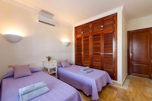 A hotel room with two beds, a wardrobe, a bedside table with a lamp, and an air conditioning unit.