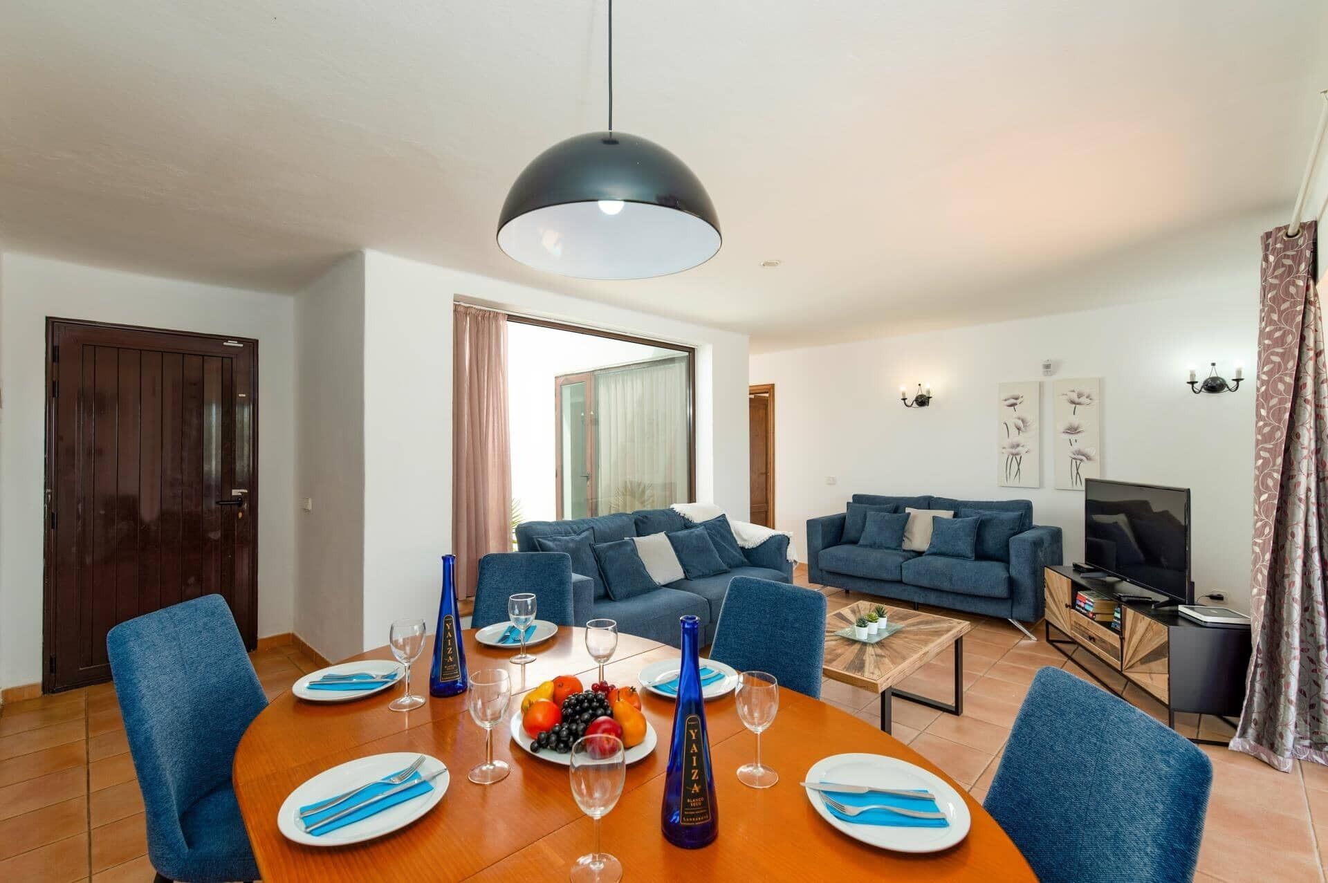 A modern living room with a dining area, featuring a large round table, blue upholstered sofas, a television, and a chandelier.