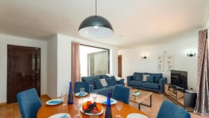 A modern living room with a dining area, featuring a large round table, blue upholstered sofas, a television, and a chandelier. - Villa Yaiza by Villa Plus (Playa Blanca)