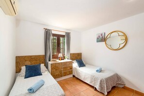 Un dormitorio con dos camas, un aparador de madera, una ventana con cortinas, un espejo redondo y un pequeño cuadro en la pared.