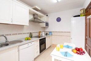 A compact kitchen with white cabinets, a microwave, a stove, and a sink. There is a dining table with plates and cutlery, and a clock on the wall. - Villa Lomo Blanco by Villa Plus (Playa Blanca)