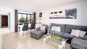 Un salon moderne avec un canapé gris, une table basse en verre et un coin repas comprenant une table et des chaises blanches.