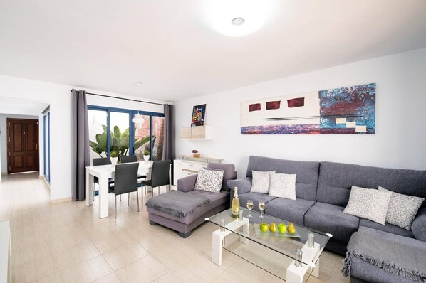 A modern living room with a grey sofa, a glass coffee table, and a dining area with a white table and chairs. - Villa Lomo Blanco by Villa Plus (Playa Blanca)