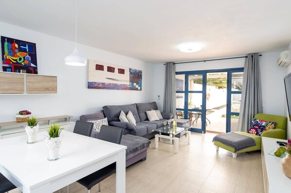 A modern living room with a grey sofa, a white dining table, and a green armchair. There is a glass coffee table and a painting on the wall.