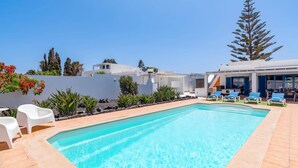 Une piscine avec des chaises longues, un bâtiment blanc et un grand arbre.