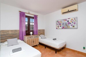 Une chambre avec deux lits, une fenêtre ornée de rideaux violets, un climatiseur fixé au mur et un tableau abstrait aux couleurs vives.
