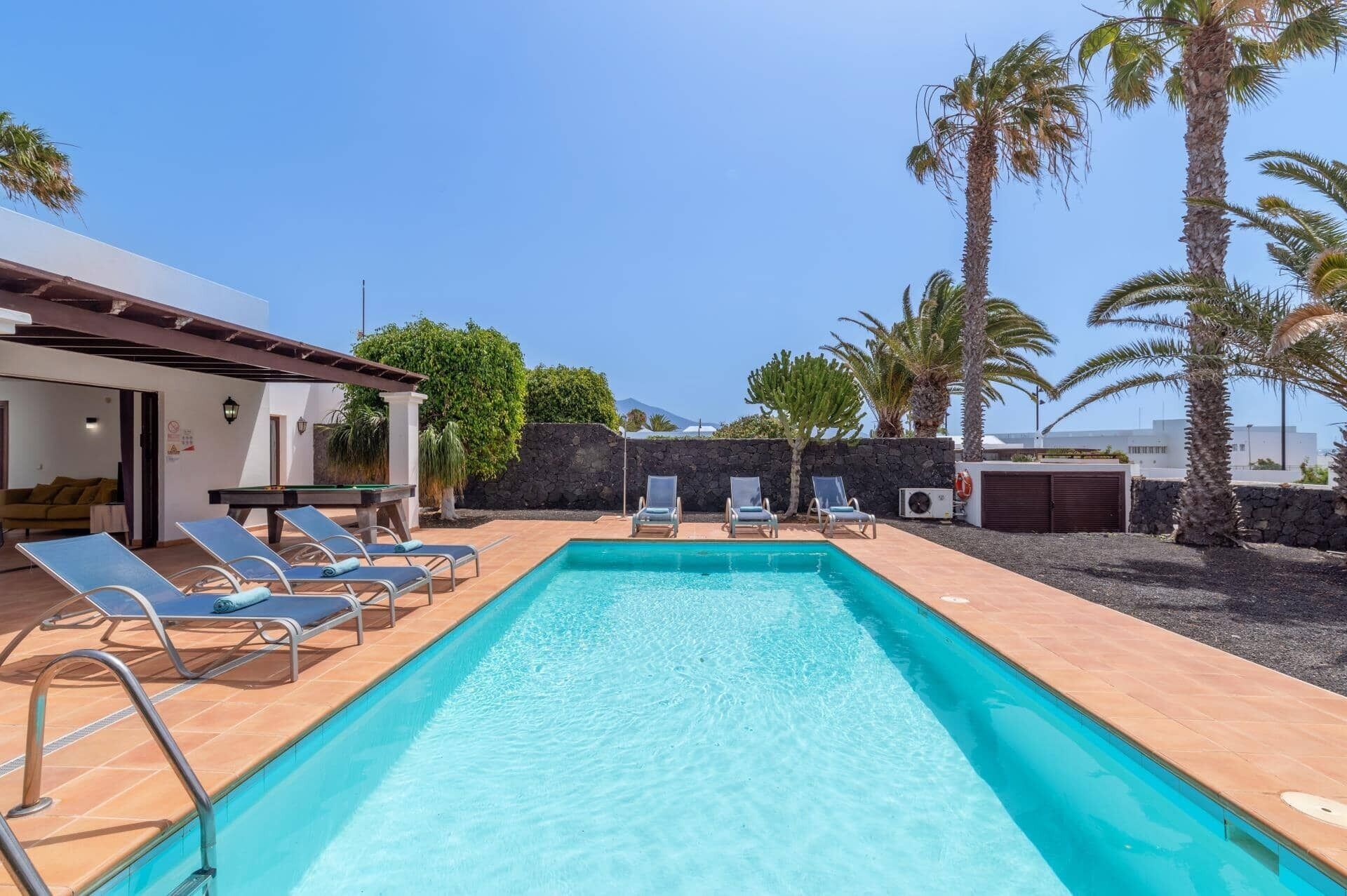 Zona de piscina con sillas de descanso, piscina y palmeras.