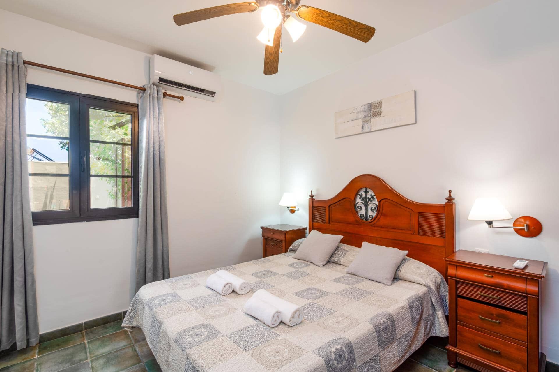 A bedroom with a bed, a nightstand, a wall-mounted lamp, a ceiling fan, and a window with curtains.