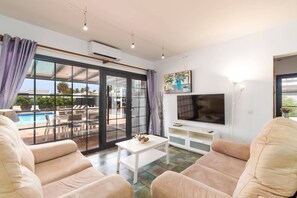 A modern living room with a large TV, comfortable seating, a coffee table, and a view of an outdoor pool area. - Villa Yeray by Villa Plus (Playa Blanca)