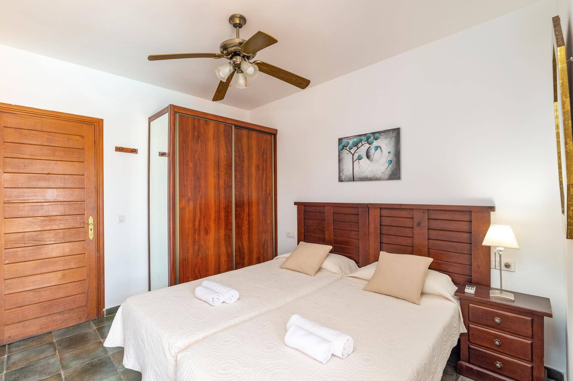 Habitación de hotel con una cama, un armario de madera, un ventilador de techo, un cuadro y una mesita de noche con lámpara.
