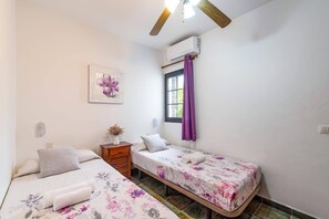 A bedroom with two beds, a window with purple curtains, an air conditioning unit, and a painting on the wall. - Villa Yeray by Villa Plus (Playa Blanca)