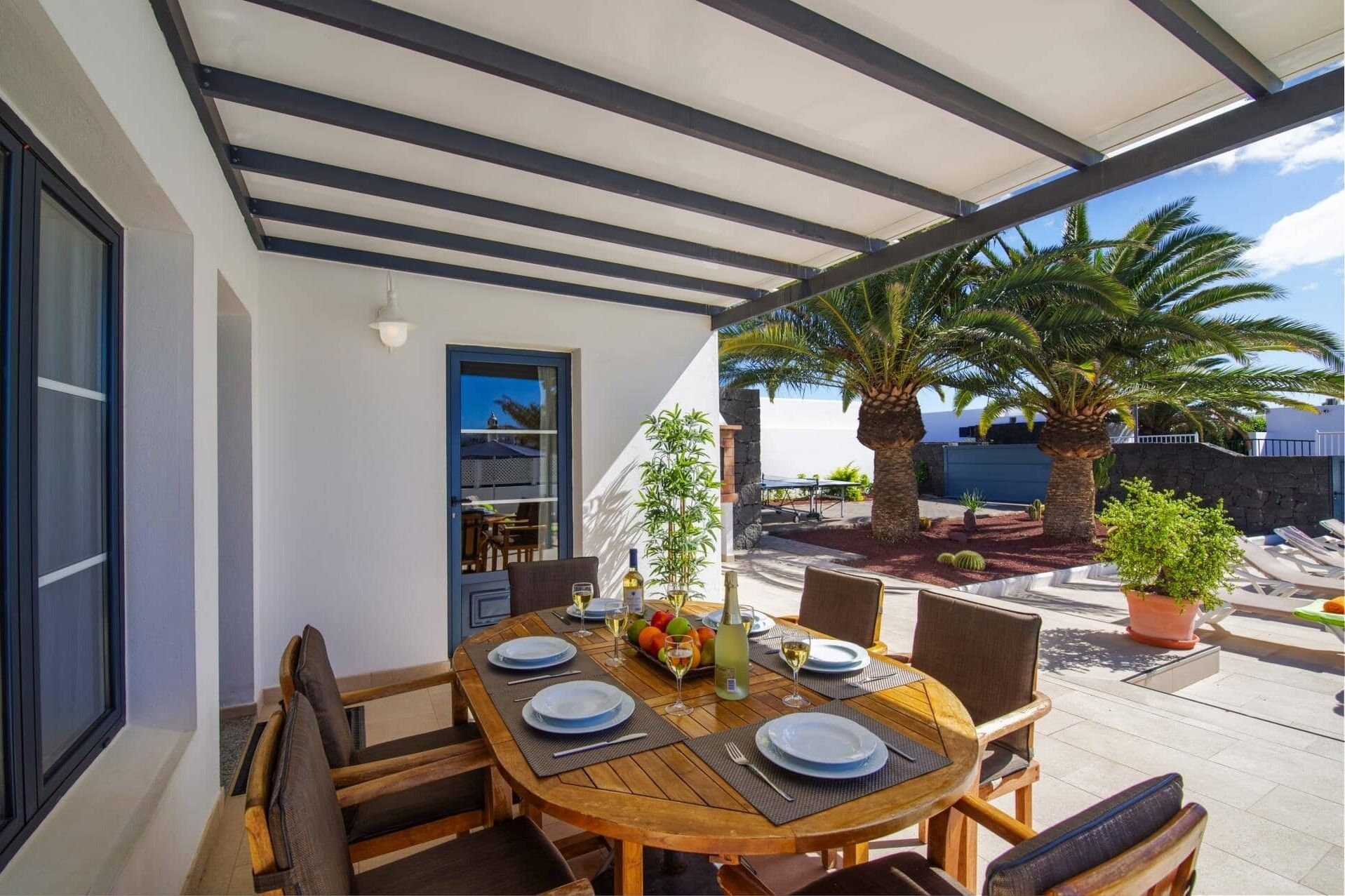 Un espace extérieur abrité, aménagé pour les repas, avec une table pour quatre personnes, entourée de chaises, et donnant sur un jardin agrémenté de palmiers et d’un mur en pierre.
