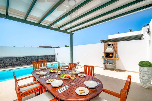 An outdoor dining area with a pool, wooden table, chairs, and a barbecue grill.