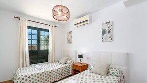 Ein Zimmer mit zwei Betten, einem Vorhangfenster, einer Pendelleuchte, einer Klimaanlage und Wandbildern.