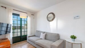 Ein modernes Wohnzimmer mit einer Couch, einem Fernseher, einem kleinen Tisch mit Pflanze und einem großen Fenster mit Vorhängen.