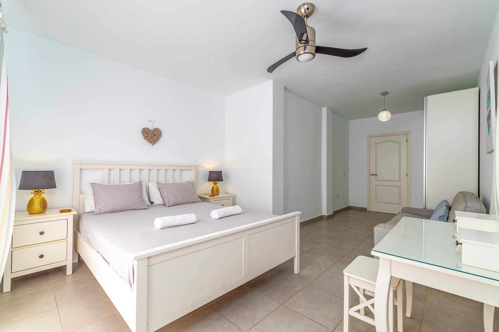 Un dormitorio con cama, mesitas de noche, ventilador de techo, una decoración con forma de corazón y una puerta que da a otra habitación.