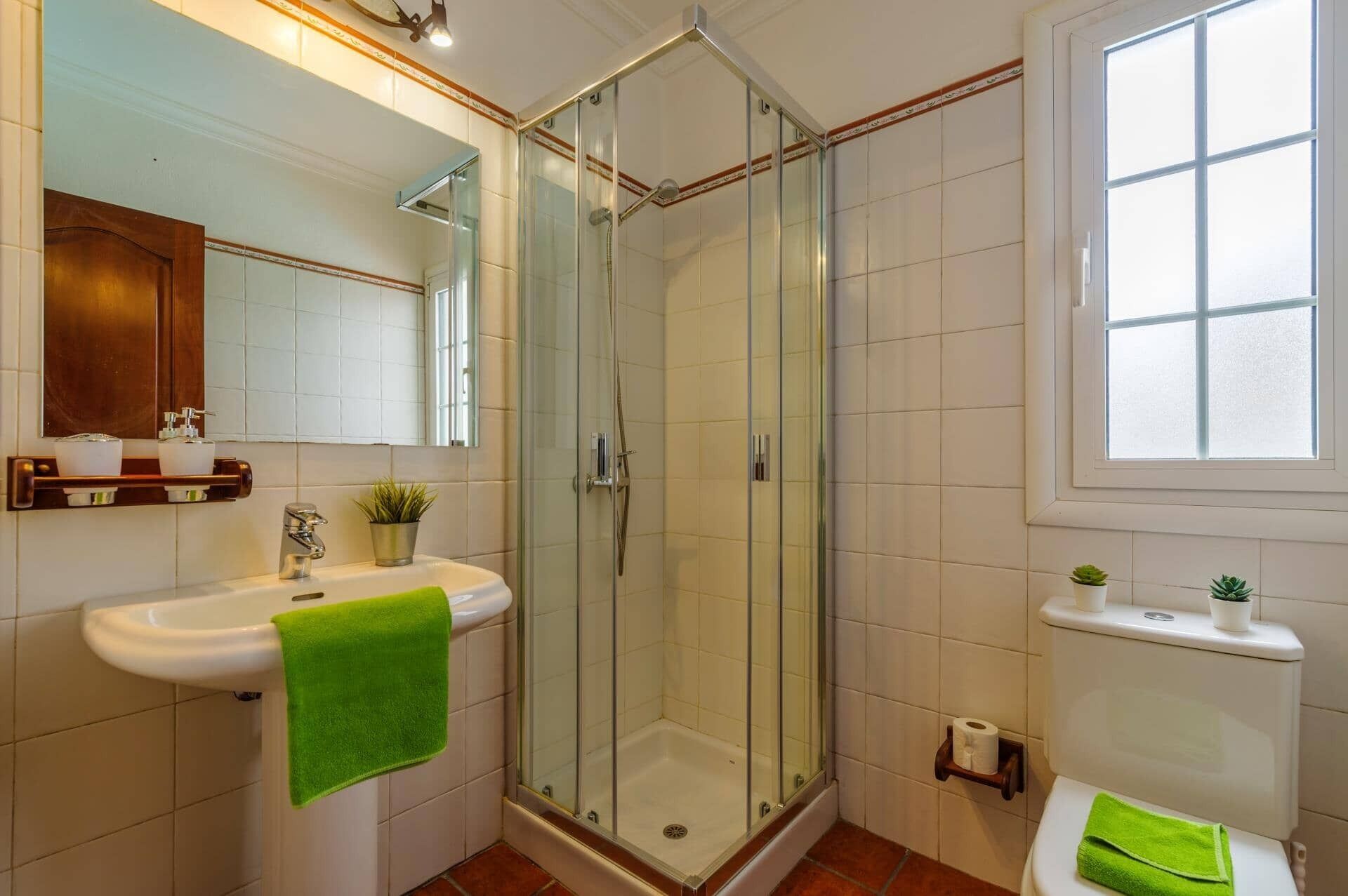 Ein Badezimmer mit einer gläsernen Duschkabine, einem Waschbecken mit einem grünen Handtuch, einer Toilette und einem Fenster.