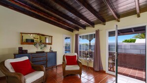 Una habitación con piso de baldosas, techo de madera, una silla de mimbre con un cojín rojo, un aparador azul y un gran ventanal con vistas.
