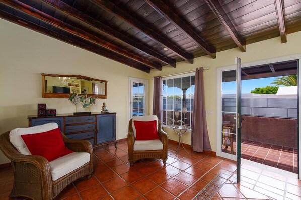 Una habitación con piso de baldosas, techo de madera, una silla de mimbre con un cojín rojo, un aparador azul y un gran ventanal con vistas.