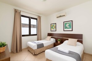 A hotel room with two beds, a window with curtains, an air conditioner, and two framed floral paintings.