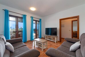 Una sala de estar con un conjunto de sofás, un televisor sobre un mueble de madera y un balcón con cortinas azules.