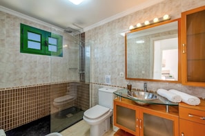 Un baño con cabina de ducha de vidrio, inodoro, lavamanos con encimera de vidrio y espejo con luces superiores.