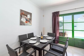Een eetgedeelte met een tafel voor vier personen, rieten stoelen, een schilderij aan de muur en een groene deur met een raam.