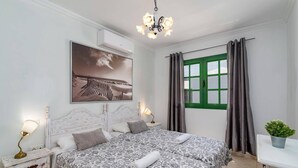 Un dormitorio con cama, mesitas de noche, lámpara, cuadro, ventana con cortinas y planta en maceta.