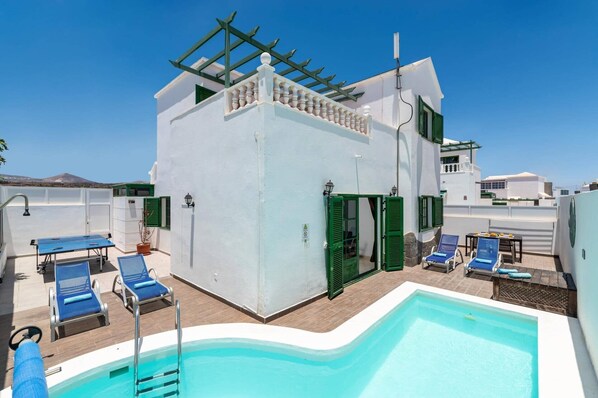 Zona de piscina en la azotea con piscina, sillas de descanso y un edificio con contraventanas verdes.