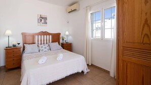 Un dormitorio con una cama de madera, dos mesitas de noche con lámparas, una ventana con cortinas y un equipo de aire acondicionado.