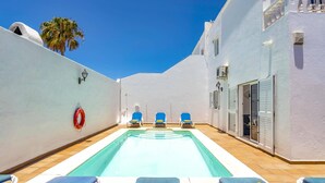 Zona de piscina con sillones reclinables azules y amarillos, un flotador salvavidas y un edificio blanco con piso de baldosas.