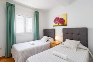 A hotel room with two beds, a window with curtains, a bedside lamp, and a painting on the wall.