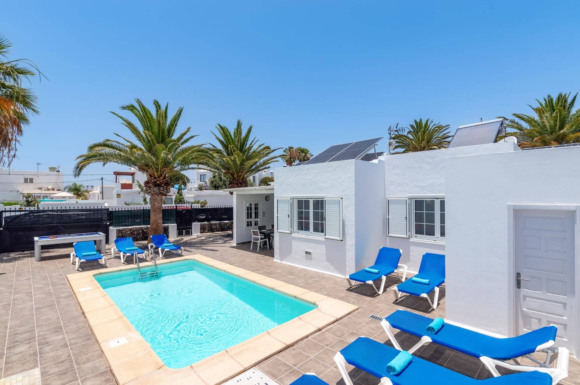 Un espace piscine avec des chaises longues bleues, un bâtiment blanc et des palmiers.
