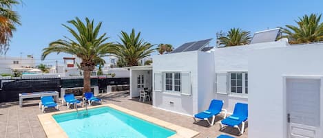 Un espace piscine avec des chaises longues bleues, un bâtiment blanc et des palmiers.