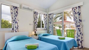 Habitación con dos camas, cada una con colcha azul acolchada y toalla verde. Cuenta con una ventana con cortinas, un equipo de aire acondicionado y vistas a palmeras desde el exterior.