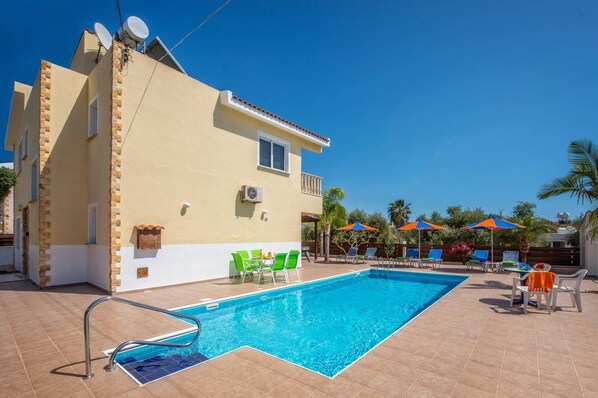 A swimming pool with a metal ladder, surrounded by lounge chairs and umbrellas, and a building with a satellite dish. - Villa Semeli by Villa Plus (Peyia)