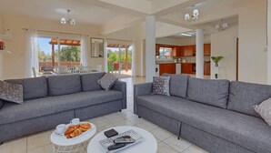 A spacious living area with two grey sofas, a coffee table, and a dining table with chairs. There is a kitchen area visible in the background. - Villa Semeli by Villa Plus (Peyia)