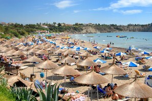 A beach with numerous sun umbrellas and lounge chairs, a rocky coastline, and a clear sky. - Villa Pandora by Villa Plus (Peyia)