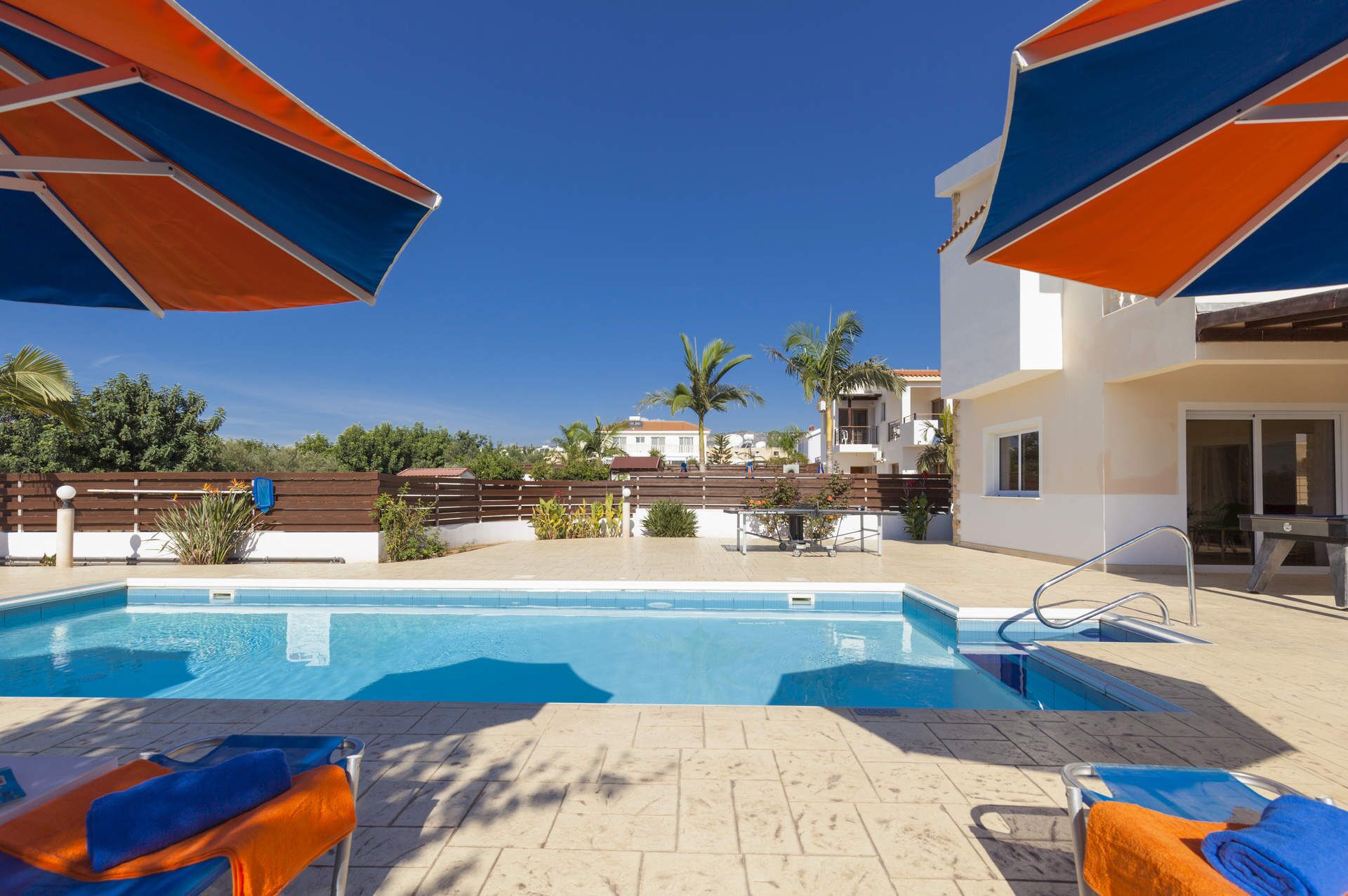 A swimming pool with lounge chairs and umbrellas, surrounded by a wooden fence and palm trees.