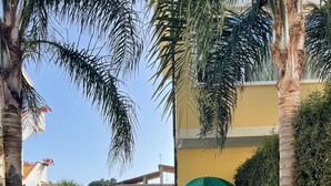 Ein Weg führt zu einem Gebäude mit gelber Fassade, umgeben von gepflegter Bepflanzung und Palmen.