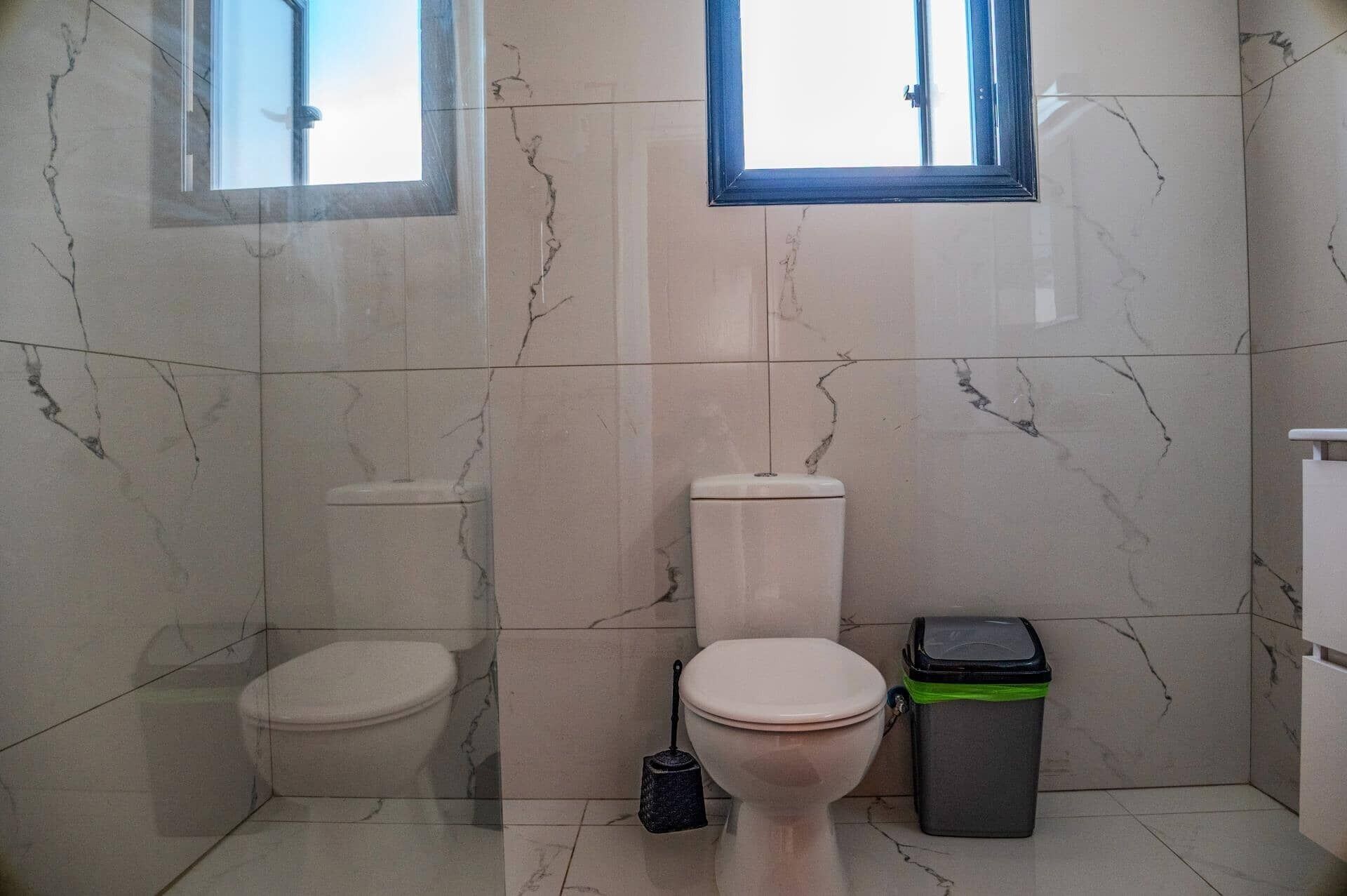 A bathroom with two toilets, a trash bin, and a window.